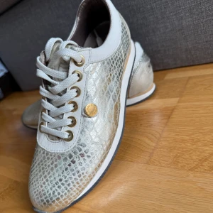 Silvriga sneakers från Novita - •Ett par snygga sneakers från Novita.  •Skorna har ett ormskinnsmönster.  •Detaljer i guld.  •Perfekt för en avslappnad men stilfull look.  •Ovandelen är i skinn med en cool glansig finish och märkets logga på hälen. •Äkta skinn. Storlek 38. Sparsamt använda. Mycket bra skick, ingenting att anmärka på. •Köpte för 1900 kr.  •Djurfritt och rökfritt hem.