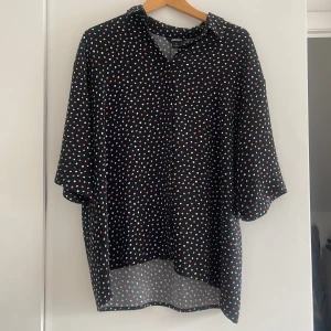 Svart prickig skjorta från Monki - Svart skjorta från Monki med färgglada prickar i vitt, rosa, turkos och orange. Skjortan har lös passform, kortare ärmar och klassisk krage. Perfekt för dig som gillar lekfulla mönster och oversized stil.