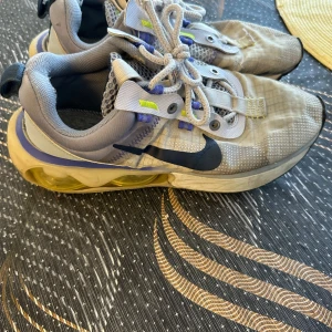 Nike sneakers med transparent sula - Nike sneakers i grått och beige med svarta detaljer och gula accenter. Skorna har en transparent, gulaktig sula med synlig dämpning och meshpartier för extra ventilation. Klassisk snörning och rund tå. Perfekta för dig som gillar sportig och modern stil.