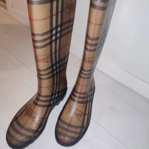 Burberry rutiga stövlar strl 37 - Snygga höga stövlar från Burberry i klassiskt beige och svart rutigt mönster. Tillverkade i syntetmaterial med platt sula och rund tå. Perfekta för regniga dagar när du vill sticka ut med en ikonisk look.