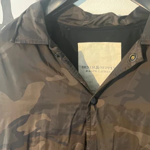 Camouflage vindjacka från Ralph Lauren - Snygg vindjacka från Denim & Supply Ralph Lauren i brunt camouflage-mönster. Jackan har klassisk krage, knäppning framtill och är tillverkad i lätt polyester. Perfekt för dig som vill ha en streetig och avslappnad look.