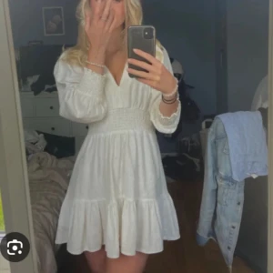 Vit kort klänning med v-ringning - Superfin vit kort klänning med v-ringning och smock i midjan. Klänningen har långa puffärmar och volang nertill, vilket ger en söt och somrig känsla. Perfekt för varma dagar och passar till sneakers eller sandaler.