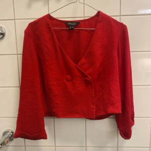 Röd croppad topp från New Look - Snygg röd croppad topp från New Look med v-ringning och två klädda knappar framtill. Toppen har lång ärm och en loose fit, perfekt för att lyfta din outfit med en pop av färg. 