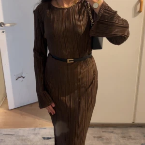 Brun plisserad långklänning med bälte - Snygg brun långklänning med plisserat tyg och lång ärm. Klänningen har ett svart bälte i midjan som ger en markerad siluett. Perfekt för dig som gillar stilrena och trendiga plagg med en twist.