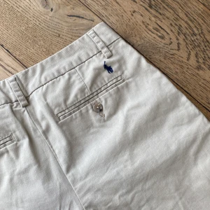 Ralph lauren chinos barn - Snygga beige chinos från Polo Ralph Lauren med klassisk rak passform. Byxorna har två bakfickor med knappar, bälteshällor och den ikoniska blå broderade loggan ovanför ena bakfickan. Tillverkade i mjuk bomull för en skön känsla. Storlek 16 vilket motsvarar 156-161 cm