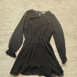 Svart kort klänning från Kappahl - Supersnygg svart klänning från Kappahl med långa transparenta ärmar, markerad midja och volangkant nertill. Perfekt för dig som gillar en enkel men ändå elegant stil. Lätt och skönt material som ger en flowy känsla.
