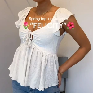 Vit blus med volang och knyt - Supersöt vit blus med volangärmar och knyt framtill. Toppen har en lätt rynkad struktur och v-ringning, samt en flowy, luftig passform som ger en romantisk vibe. Perfekt till jeans eller kjol för en fräsch look.