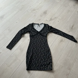 Svart blommig kortklänning H&M Divided - Supersöt svart kortklänning från H&M Divided med små vita och rosa blommor. Klänningen har lång ärm, v-ringning med spetsdetalj och figurnära passform. Perfekt för dig som gillar en feminin och trendig stil!