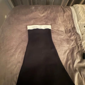Svart långklänning med vit kant - En elegant svart långklänning med en bred vit kant upptill. Klänningen har en figurnära passform och är axelbandslös, vilket ger en stilren och modern look. Perfekt för dig som vill sticka ut med en klassisk men trendig stil.