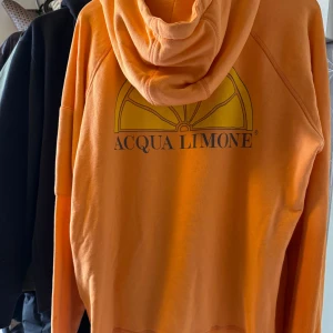Orange hoodie från Acqua Limone str L - Säljer en orange hoodie från Acqua Limone med klassisk citronskiva och logga tryckt på bröstet och ryggen. Tröjan har huva med snörning, lång ärm och en ficka med dragkedja på ena ärmen. Mjuk och skön bomullskvalitet, strl L. Aldrig använd 