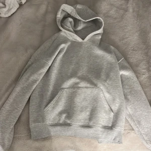 Ljusgrå hoodie från Gina Tricot - En basic ljusgrå hoodie från Gina Tricot med stor känguruficka framtill och mjuk insida. Tröjan har huva, långa ärmar och ribbade muddar vid ärmslut och nederkant. 