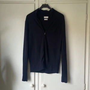 Svart merinoullströja från These Glory Days - Svart långärmad tröja från These Glory Days i mjuk och lyxig merinoull. Tröjan har en stilren design med half full-dragkedja framtill och ribbade muddar. Perfekt för dig som gillar minimalistisk och clean stil.