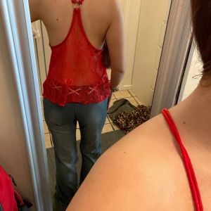 Röd linnetopp med spetsdetaljer - Säljer en röd linnetopp med tunna axelband och v-ringning framtill. Toppen har en snygg blandning av glansigt tyg framtill och transparent spets med blommönster baktill. Perfekt för dig som vill sticka ut med en färgstark och feminin look.