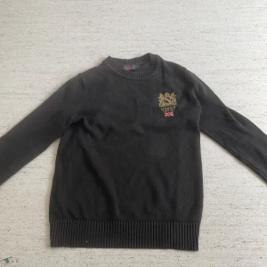 Mörkgrön stickad tröja från Morris XS - Mörkgrön stickad tröja från Morris med broderad guldfärgad logga och brittisk flagga på bröstet. Klassisk rund halsringning och ribbade muddar vid ärmslut och nederkant. Perfekt för dig som gillar stilren och tidlös design.  Storlek xs men passar S fint, då jag uppfattade det som att den var stor i sin storlek