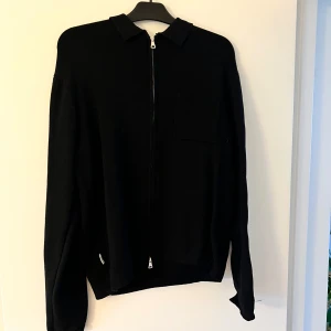 Svart stickad overshirt Studio Total - Svart stickad overshirt från Studio Total i storlek M. Jackan har klassisk krage, dragkedja framtill och en bröstficka. Perfekt för lager-på-lager och enkel att matcha med allt. Långärmad och i mjukt material som känns skönt mot huden.