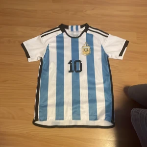 Argentina Messi #10 fotbollströja - Säljer en klassisk Argentina fotbollströja från world cup 2022 med blå och vita ränder, svart nummer 10 på bröstet och ryggen samt Messi-tryck. Broderat AFA-emblem med tre stjärnor på bröstet. Kortärmad modell i lätt och ventilerande material, perfekt för fotbollsälskare. Har använt 2 gånger 