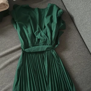 Grön omlottklänning med plisserad kjol - Snygg lång grön klänning med omlottliv och plisserad kjol. Klänningen har korta ärmar och ett knytband i midjan som ger en fin siluett. Perfekt för dig som vill sticka ut med en elegant och trendig look.
