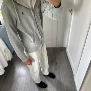 Grå zip från Polo Ralph Lauren - Grå hoodiejacka från Polo Ralph Lauren med dragkedja, huva med vita snören och två fickor framtill. Klassisk broderad logga på bröstet. Mjuk bomullskänsla och perfekt för chill dagar eller när du vill ha en clean look.