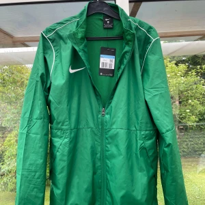 Grön Nike vindjacka med dragkedja - Snygg grön vindjacka från Nike med vit logga på bröstet och tunna vita detaljer längs axlarna. Jackan har hel dragkedja framtill, ståkrage och är tillverkad i lätt polyester. Perfekt för dig som gillar sportig stil och vill ha något lätt att slänga på sig. Använd 1 gång och säljer då den är för liten för mig, helt ny skick med lappen kvar.