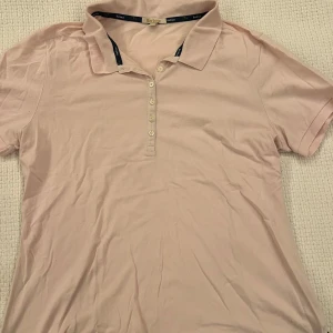 Ljusrosa piké från Barbour - Säljer en ljusrosa pikétröja från Barbour med klassisk krage och knappslå med flera knappar. T-shirten är kortärmad och har en liten metallbricka med Barbour-logga. Skön bomullskvalitet och perfekt för en clean och stilren look.