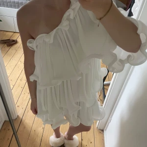Vit oneshoulder volangklänning från nakd - Superfin vit kortklänning från nakd med oneshoulder och stora volangdetaljer. Klänningen har plisserat tyg och en luftig, flowy passform som känns riktigt trendig. Perfekt för sommarens alla tillfällen när du vill sticka ut lite extra.