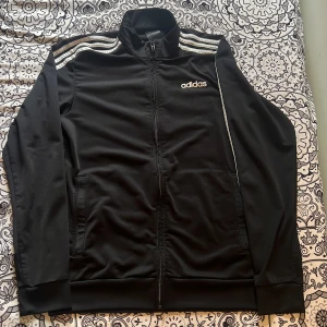 Svart Adidas track jacket med stripes - Svart Adidas track jacket med klassiska vita stripes på axlarna och dragkedja hela vägen. Jackan har hög krage, två fickor och Adidas-logga på bröstet. Perfekt för en sportig och avslappnad stil.