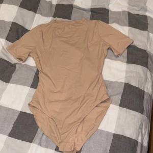 Beige kortärmad bodysuit från skims  - En stilren beige bodysuit med korta ärmar och rund hals. Bodyn har knäppning i grenen för enkel på- och avtagning. Perfekt att styla med jeans eller kjol för en clean och enkel look.