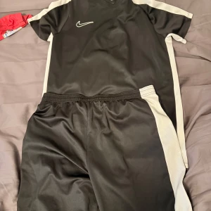 Svart Nike avademy dri fit T-shirt set - Snygg svart fotbollströja från Nike i Dri-FIT-material som andas. Tröjan har korta ärmar och vita paneler längs sidorna och axlarna. Perfekt för träning eller match, med klassisk Nike Swoosh på bröstet. Blivit för små för mig.