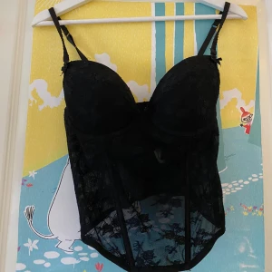 Svart spetskorsett med justerbara band - Säljer en svart korsett med spetsdetaljer och vadderade kupor. Toppen har justerbara axelband och hakknäppning i ryggen. Transparent mesh och blommig spets ger en snygg look. Perfekt att styla med jeans eller kjol för en trendig outfit.