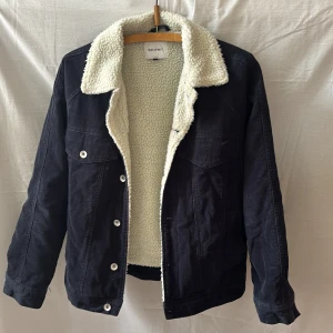 Svart teddyjacka från Vailent - Svart teddyjacka från Vailent i manchester med vit, fluffig teddyfoder på insidan och kragen. Klassisk jeansjacke-look med knappar framtill och två bröstfickor. Perfekt för kyliga dagar och ger en cool, avslappnad vibe.