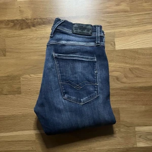 Replay Anbass blå jeans med slitningar - Säljer ett par Replay Anbass jeans i klassisk blå tvätt med snygga slitningar över knäna. Jeansen har fem fickor, smal passform och tydliga Replay-detaljer på bakfickan. Perfekta för dig som gillar en modern och avslappnad look. Tyvärr är storleksettiketek borta så kolla måtten noggrant men den tidigare ägaren hävdar att det är W31 L30 och jag tycker att det stämmer ;)