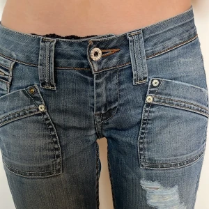 Lågmidjade Blå jeans med detaljerade bakfickor - Säljer ett par blå jeans med slitningar framtill och unika bakfickor med dragkedjor, nitar och färgade ränder i brunt, orange och vitt. Jeansen har raka ben, flera fickor och coola sömdetaljer. Perfekta för dig som gillar en streetig och personlig stil.