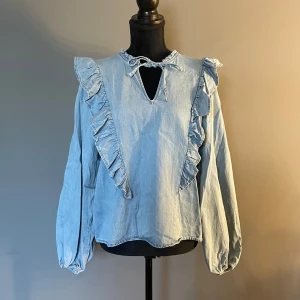 Blå blus från Kappahl - Denna fina blå blus från Kappahl, ser ut att vara i jeans material. Den har volanger fram och baktill. Fram på blusen finns 2 band man kan välja att låta hänga eller knyta för en extra ditalj.