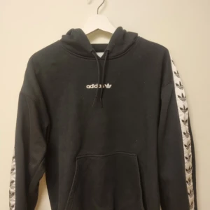 Svart Adidas hoodie med logga S - Svart hoodie från Adidas med vit broderad logga på bröstet och vita band med svarta Adidas-loggor längs ärmarna. Klassisk känguruficka framtill, huva med snörning och mjukt bomullsmaterial. Perfekt för en sportig och avslappnad stil.