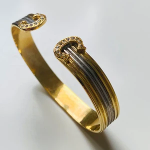 Guldigt armband med hästskor och stenar - Unikt armband i guld- och silverfärgad metall med räfflade linjer och öppna ändar dekorerade med gnistrande stenar i form av hästskor. Perfekt för dig som gillar detaljer med hästinspiration och vill sticka ut med en cool accessoar.