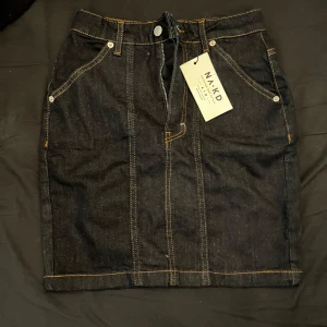 Svart jeanskjol från NA-KD - Snygg svart jeanskjol från NA-KD med klassiska fickor fram och bak, synliga sömmar i kontrastfärg och bälteshällor. Kjolen har dragkedja och knapp framtill och är perfekt för en trendig vardagslook.