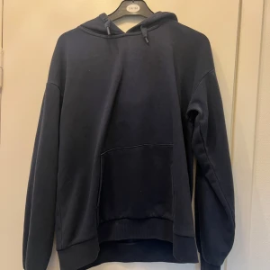 Mörkblå hoodie med huva - Enkel och stilren mörkblå hoodie med huva och känguruficka framtill. Tröjan har långa ärmar och ribbade muddar vid ärmslut och nederkant. Perfekt för dig som gillar en avslappnad och bekväm stil.