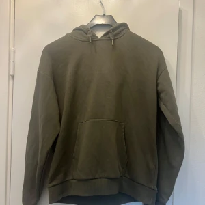 Olivgrön hoodie med ficka - Säljer en olivgrön hoodie med huva och snörning. Tröjan har en stor magficka och ribbade muddar vid ärmslut och nederkant. Perfekt för dig som gillar en avslappnad och enkel stil. Passar både tjejer och killar.