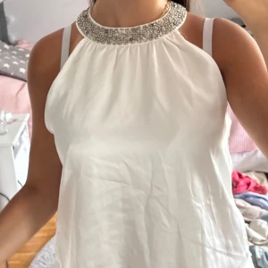 Vit halterneck topp med pärldetaljer - Supersnygg vit halterneck topp i glansigt material med bred, dekorerad krage med silvriga pärlor.
