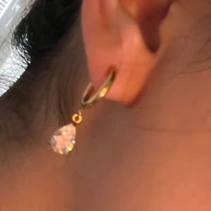 Guldörhänge med droppformad sten - Snyggt örhänge i guldton med droppformad klar sten som hänger från en tunn ring. Perfekt för dig som gillar stilrena smycken med lite extra bling. Stenen fångar ljuset superfint och ger en lyxig känsla.