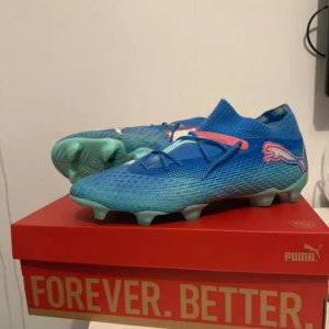 Blåa Puma fotbollsskor med snörning - Snygga blåa fotbollsskor från Puma med stickad ovandel och vita loggor på sidorna. Skorna har snörning, rosa detaljer och en strumpliknande passform för extra komfort. Sulan är ljusgrön och anpassad för spel på gräs. Perfekta för dig som vill sticka ut på planen.