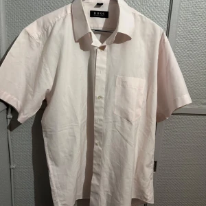 Ljusrosa kortärmad skjorta från BOSS - Snygg ljusrosa skjorta från BOSS med korta ärmar och klassisk krage. Skjortan har en bröstficka med diskret broderad logga och stängs med knappar framtill. Perfekt för en clean och stilren look.