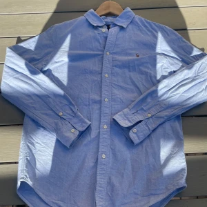 Blå skjorta från Ralph Lauren - Klassisk ljusblå skjorta från Ralph Lauren med broderad logga på bröstet. Skjortan har lång ärm, knappar framtill och är tillverkad i bomull. Perfekt för en clean och stilren look.
