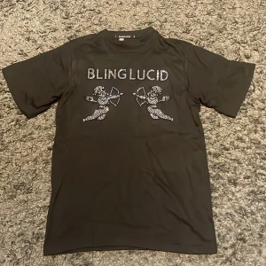 Svart t-shirt med strasstenar Blinglucid - Svart t-shirt från Blinglucid med coola strasstenar framtill som bildar texten BLINGLUCID och två cupider. Klassisk passform och korta ärmar, perfekt för dig som gillar statement-plagg med bling.