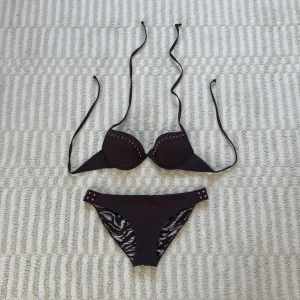 Brun bikini med nitar  - Snygg brun bikini från H&M med lätt push-up kupa och dekorativa kopparfärgade nitar på både överdel och underdel. Insidan har zebramönstrat tyg i svart, beige och brunt. Bikinin har knytband i nacken och klassisk passform. Perfekt för stranden eller poolen!