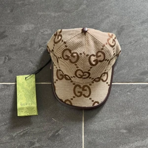 Beige keps från Gucci med GG-logga jumbo.  - Snygg beige keps från Gucci med klassiskt brunt GG-monogram över hela kepsen. Kepsen har bruna detaljer och justerbar rem bak. Tillverkad i Italien och i ett tåligt bomullsmaterial. Perfekt för dig som vill sticka ut med en lyxig accessoar.