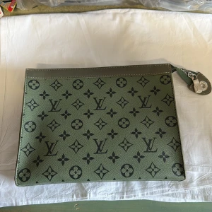 Grön Louis Vuitton kuvertväska - Snygg grön kuvertväska från Louis Vuitton med klassiskt monogrammönster i svart. Väskan har silverfärgad dragkedja och detaljer, samt praktisk handledsrem. Tillverkad i skinn med exklusiv känsla och röd insida. Perfekt för dig som vill sticka ut med stil.