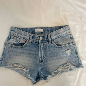 Blå jeansshorts från Gina Tricot - Snygga ljusblå jeansshorts från Gina Tricot med råa kanter och slitna detaljer. Klassisk femficksmodell med knapp och dragkedja framtill. Perfekta för sommaren och ger en avslappnad vibe till din outfit.