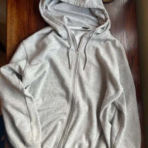 Grå zip-up hoodie Gina Tricot - Mjuk och basic ljusgrå hoodie från Gina Tricot med hel dragkedja framtill, huva med snörning och långa ärmar. Tillverkad i bomullsmix med borstad insida för extra komfort. Perfekt för chill dagar eller när du vill ha en enkel och clean look.