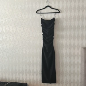 Svart långklänning med tunna axelband - Supersnygg svart klänning till nästan ankeln med tunna axelband och draperad front. Klänningen har en tight passform och är perfekt för dig som vill ha en elegant och stilren look. Materialet känns mjukt och följsamt mot kroppen.